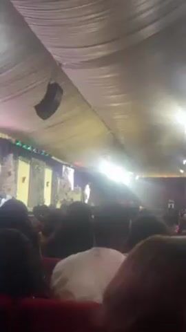 The video shows an indoor setting, possibly a church or auditorium, filled with people. The camera is positioned from the back of the room, looking towards a stage. A speaker is visible on the stage, addressing the crowd. The room is well-lit with overhead lights. The ceiling is covered with white fabric, giving a tent-like appearance. A large speaker is suspended from the ceiling on the left side of the frame. The people in the audience are of various ages and genders, with their backs facing the camera. The overall atmosphere appears to be attentive and engaged, with the audience focusing on the speaker on stage.