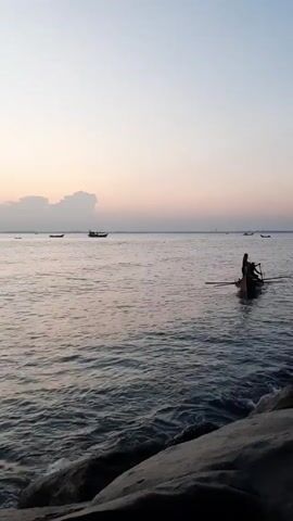 The video shows a wide shot of a body of water at dusk. Several small boats can be seen on the water, including one close to the shore. This boat contains three people, two of whom appear to be rowing. The sky is a pale gradient of light blue to light orange, and a line of clouds are visible on the horizon. In the foreground, there are several large rocks. The water is choppy.