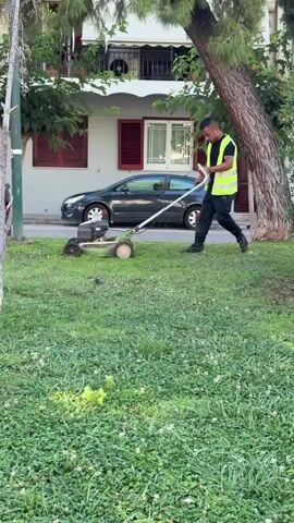 A man wearing a bright yellow safety vest over a black t-shirt and black pants is operating a lawnmower. He is pushing the lawnmower forward across a patch of grass. The lawnmower is silver and green. Behind the man, there is a building with balconies and a parked car. There is also a tree with green foliage. The ground is covered in grass and some clover. The scene appears to be an outdoor urban environment.