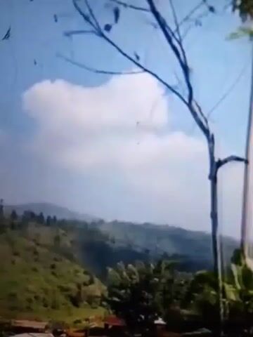 The video shows a scenic view of rolling hills covered in lush green vegetation under a bright blue sky with scattered white clouds. In the foreground, a few bare branches of a tree and some green leaves are visible. In the middle ground, a small cluster of houses with reddish-brown roofs is nestled among the hills. The overall impression is one of a peaceful rural landscape.