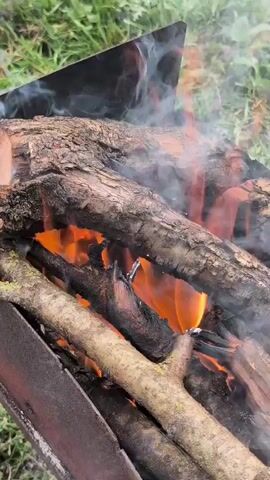The video shows a close-up of a campfire. Logs of various sizes are stacked in a metal fire pit. Flames and smoke rise from the wood, indicating it is burning. The background is out of focus but appears to be a grassy, outdoor environment. There are no people visible in the video.