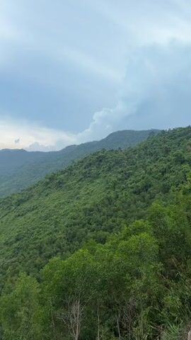 The video shows a landscape of lush, green mountains under a sky that transitions from cloudy and overcast to partly cloudy with patches of blue. The camera slowly pans upwards, revealing a series of mountain ranges stretching into the distance, their slopes densely covered in trees. In the valley below, a glimpse of civilization can be seen, possibly a village or town.