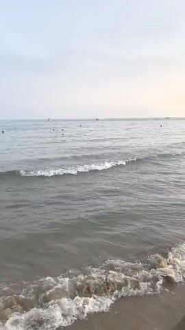 The video shows a calm ocean scene with gentle waves lapping at a sandy shore. Several people are visible in the shallow water, appearing to be swimming or wading. In the distance, a few boats are also present. The sky is overcast and the light suggests it may be early morning or late evening. The overall atmosphere is peaceful and serene.