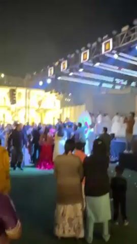 The video shows an outdoor event, likely a wedding reception, taking place at night. There's a crowd of people gathered, many appearing to be dancing or moving to the music. Some people are wearing traditional clothing, including a woman in a red and gold dress and others in saris. There are also people wearing Western clothing. In the background, there's a large stage-like structure with lighting and what looks like a facade resembling a building. The ground appears to be covered with artificial turf. The camera moves slightly and sometimes shakes, suggesting it's being held by someone moving through the crowd.