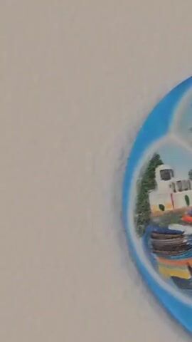 The video shows a tourist souvenir plate hanging on a wall. The plate is round and blue, with the word "POMORIE" written at the top. The plate is divided into four sections, each depicting a different scene from the town. In the top section, there is a painting of a modern building next to water. The next section down features a white building with some boats in the water. Next to that is a scene of a historic-looking stone building. The final section depicts some buildings with water in the background. In the center of the plate, there is a raised decoration made of colorful shapes.