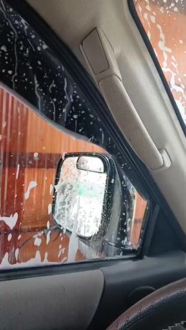 The video shows the interior of a vehicle during a car wash. Soap suds are visible on the window and the side mirror. Through the mirror, it is possible to see a blurry green object. The background consists of an orange ribbed wall, likely part of the car wash facility. The timestamp indicates a duration of 5 seconds.
