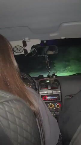 The video shows the interior of a car at night. The camera is positioned in the back seat, looking forward. In the foreground, the long brown hair of a person is visible. In the middle ground, a woman with dark hair is sitting in the passenger seat, wearing a gray sweatshirt. She turns to the camera and smiles, making a rock-and-roll hand gesture. The dashboard of the car is visible, with a radio and air vents. Outside the car, the headlights illuminate a dark, possibly rural road. The scene appears to be a candid moment captured while driving.