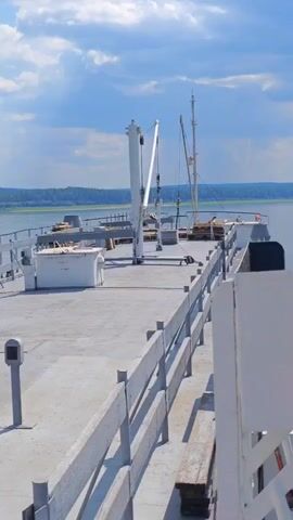 The video pans across the deck of a ship, showing two cranes and some cargo. The sky is blue with white clouds, and there is land in the distance. The deck is gray and has a railing along the side.