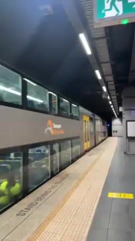 A double-decker train, identified as "Transport Sydney Trains," is stopped at an underground train station. The train is gray with yellow doors, and its windows show interior seating. The station platform is marked with a textured yellow tile and the words "STAND BEHIND THE YELLOW LINE" in black lettering. The station ceiling is dark, with overhead fluorescent lights. An emergency exit sign with a green running man is visible in the upper right corner. The scene appears clean and well-maintained, with a security camera and signage also visible. The camera is handheld, and the perspective is from someone standing on the platform, stationary.