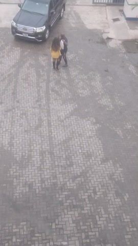 The video shows an aerial view of a paved outdoor area. A black SUV is parked on the left side of the frame. Three individuals are standing on the paved area. One person, wearing a dark top and jeans, is bending over. Another person, wearing a yellow top, is standing upright. A third person, wearing a blue top and jeans, is also standing upright. The paved area appears to be wet in places, with streaks and patterns visible on the surface. The lighting suggests it is daytime, but the sky is overcast.
