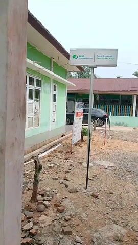 The video shows an outdoor scene with several buildings and vegetation. In the foreground, there is a gravelly and rocky area. A sign with the BPJS Ketenagakerjaan logo and text "Pusat Layanan Kecamatan" is visible, attached to a metal pole. A dark-colored car is parked in front of one of the buildings. The buildings appear to be painted green and have white window frames. Another building with a red roof is visible in the background. The overall environment seems to be a semi-rural or suburban area with trees and foliage. The ground is mostly unpaved, consisting of dirt and gravel. There are several poles and some debris scattered around. The sky is overcast. The camera appears to be handheld, moving slightly as it captures the scene.