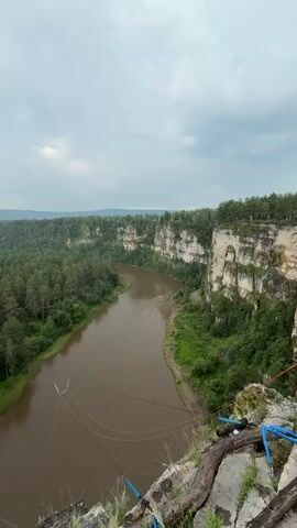 The video shows a wide, brown river flowing through a dense, green forest. The riverbanks are lined with trees, and steep, rocky cliffs rise on the right side of the river. The sky is cloudy and overcast. In the foreground, there is a wooden platform extending out over the cliff edge, with wires attached to it, suggesting it might be used for bungee jumping or a similar activity. There are no people visible in the video. The overall scene is one of natural beauty and adventure.