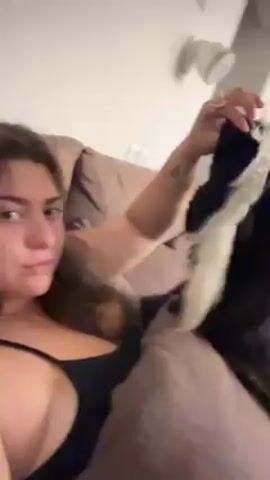 Here's a description of the video:
A woman is sitting on a couch, holding a black and white toy in front of her. The toy seems to resemble a skunk. The woman looks at the camera, playing with the toy. The couch is brown and there's a white wall behind the couch.  A speaker is mounted on the wall. The room is brightly lit.