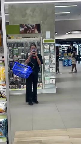 A person is standing in a store, reflected in a large mirror. They are wearing a black t-shirt and black pants and holding a blue shopping basket. The store appears to be a pharmacy or a beauty supply store, with shelves stocked with various products. In the reflection, other shoppers can be seen in the background. One person is walking away from the camera, and another is looking at products on a shelf. The lighting is bright, and the store seems clean and organized.