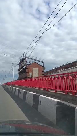 The video shows a view from a moving vehicle on a bridge. The bridge has a red railing and concrete barriers with black and white stripes. On the right side of the bridge, there is a brick building with scaffolding and electrical equipment. There are overhead power lines and a street light. In the distance, a white bus is visible driving on the bridge. The sky is cloudy.