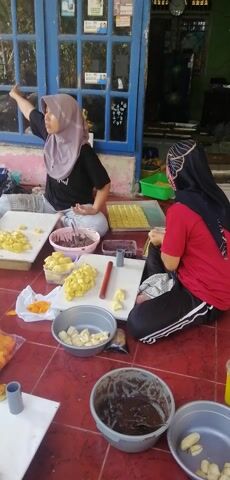 The video shows two individuals preparing food in an outdoor setting with a red tiled floor and a blue-framed window. A woman wearing a light purple hijab and a black t-shirt is on the left, cutting yellow fruit pieces on a white board. To her right, another person wearing a red shirt and black pants with white stripes is also working at a white board, appearing to use a red rolling pin.

Several containers are visible: a grey bucket on the right contains a dark brown, chocolate-like substance. A pink bucket near the center also holds a similar dark substance. A larger grey bucket on the far right contains several pieces of the same yellow fruit. A small clear container with a purple lid is also present.

Yellow fruit pieces are spread across the white boards and in a grey bowl. Bags of orange chips are on the floor to the right of the main workspace. A green tray with numerous small yellow rectangular items, possibly candy or pastries, is behind the person in the red shirt. Two grey PVC pipes are also visible. The overall environment suggests food preparation or a small food stall.