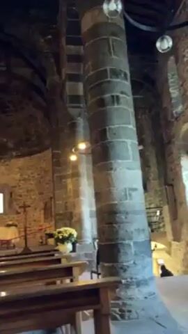 The video shows the interior of a church, focusing on the altar area. The church is built of dark stone, with large, cylindrical columns supporting vaulted ceilings. Rows of wooden pews line the nave, leading toward the altar, which is adorned with white flowers and a lace tablecloth. A blue cloth is draped to the side, near a statue of the Virgin Mary and child. The church is dimly lit by overhead lights, creating a solemn atmosphere. The camera pans slightly, showing the architectural details and religious elements.