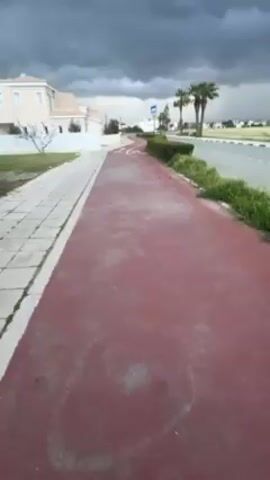 The video shows a landscape under a cloudy, overcast sky. The camera is positioned along a path that appears to be a sidewalk alongside a red-colored lane, possibly a bike path. A building is visible to the left, a light-colored, two-story structure with a white fence surrounding it. In front of the building is a grassy area and a silver car parked under a tree. Beyond the bike path, there is a road with greenery and a few palm trees on the other side. The sky is filled with heavy, dark clouds suggesting the potential for rain. The camera appears to be in motion as it moves along the path.