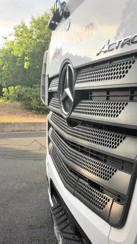 The video is a close-up, side-view shot of the front grille of a white Mercedes-Benz Actros truck. The camera pans to the left, revealing a grassy area with trees and a cloudy sky in the background. The sun is visible through the clouds, casting a warm glow on the scene. The truck is parked on an asphalt road, and a concrete barrier separates the road from the grassy area. There are no people or other vehicles visible in the video.