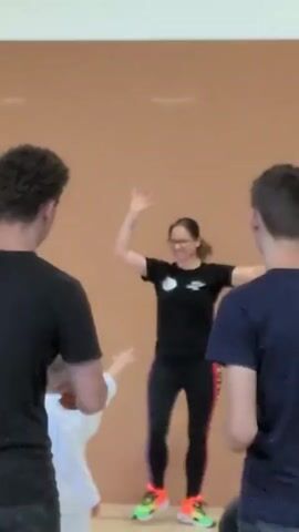 The video captures a woman in a black t-shirt and colorful leggings leading a group of children in what appears to be a dance or exercise class. She is in the center, with her arms raised and in motion, smiling. She wears glasses and her hair is pulled back. Two children in white uniforms, possibly martial arts gis, are positioned in front of her, facing away from the camera. They are also in motion, with one child's arms slightly raised. Two young men, one on the left and one on the right, are partially visible in the foreground, facing the woman and children. They are also wearing dark t-shirts. The setting is an indoor space with a light-colored wall and a light-colored floor marked with blue lines. The overall lighting is bright.