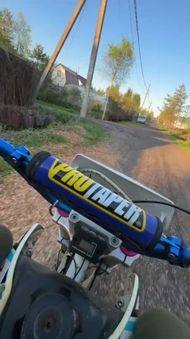 A first-person perspective video shows someone riding a dirt bike on a rural road. The camera is mounted on the bike, looking forward over the handlebars, which have blue accents and a yellow and blue ProTaper pad. The fuel tank is visible in the foreground, and a digital display is mounted near the handlebars. The road is paved but appears somewhat rough. On either side of the road are grassy areas, trees, and utility poles with wires. A few houses and a parked silver car are visible in the background. The sky is clear and blue. The rider is wearing tan pants. The bike moves steadily down the road.