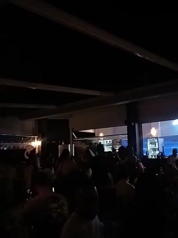 The video shows a dark, crowded indoor space, likely a bar or club, with people gathered. The lighting is dim, with some ambient light coming from behind a reflective surface on the left and from lights above on the right, possibly illuminating bottles on shelves. A ceiling structure with visible beams is overhead. The majority of the people are silhouetted and indistinct due to the low light, making it difficult to discern specific features or activities. Some individuals appear to be standing and interacting, while others are seated. The overall atmosphere is that of a social gathering in a dimly lit venue.