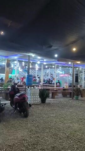 The camera pans from left to right, showing the exterior of a café at night. Several motorbikes are parked in the gravel parking area. There are wooden picnic tables, plants in pots, and lights strung above. People are visible inside the café through glass windows.  A person walks from left to right across the parking area. The camera stops on a woman sitting at an outdoor table, facing away from the camera.