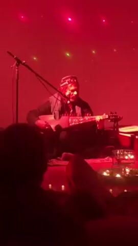 In a dimly lit room, washed in red light, a musician sits cross-legged on a rug, playing a stringed instrument. A microphone stands before him, suggesting a performance setting. The musician wears a patterned cap and dark clothing. In the foreground, a blurry audience member is visible, indicating the presence of other people.  A table sits near the musician, adorned with candles and a glass of water. The red lighting creates a warm and intimate atmosphere.