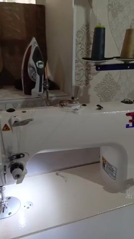 A sewing machine is being used to stitch a patterned fabric. The sewing machine is white and has a light illuminating the needle area. A spool of thread is visible on top of the machine. In the background, an iron is resting on a surface. The environment appears to be a room with wallpaper and a shelf holding additional spools of thread.