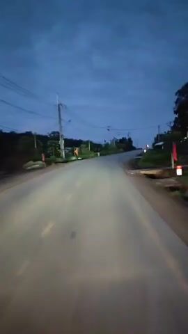 The video shows a first-person perspective of traveling on a paved road at dusk or night. The road is lit by headlights, casting a bright beam ahead and illuminating the asphalt surface, which has yellow dividing lines. Utility poles with power lines are visible on either side of the road. Vegetation, including trees and bushes, lines the sides of the road. In the distance, the road appears to curve upwards and the horizon is visible under a dark, cloudy sky. Several road signs are also present, including a yellow triangular warning sign with a black symbol. The overall environment suggests a rural or semi-rural setting.