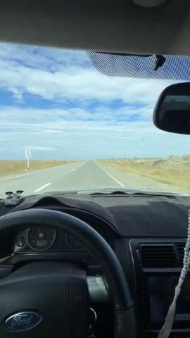 The video shows the interior of a car driving on a straight, paved road through a flat, open landscape under a cloudy blue sky. The dashboard and steering wheel are visible, with the car's instruments showing a speed of 70 km/h and the Ford logo on the steering wheel. The road stretches ahead into the distance, bordered by dry, brown fields. The sun appears to be shining from the right side of the frame, casting shadows on the dashboard. The overall impression is one of a steady, uneventful drive through a rural or steppe environment.