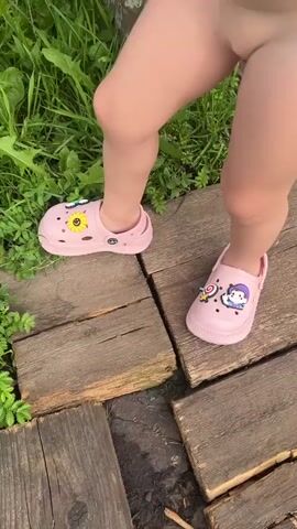 The video shows a close-up of a child's legs and feet wearing pink Crocs with cartoon charms. The child is standing on a wooden path surrounded by lush green grass and vegetation. The camera pans slightly to show the child's feet moving through the grass.