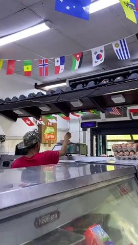 The video shows the interior of a store, likely a small grocery or butcher shop, decorated with numerous small flags hanging from the ceiling, representing various countries. A person wearing a red shirt and a camouflage cap is standing behind a counter, operating a scale or a cash register. They appear to be interacting with a customer, though the customer is not visible in the frame. In the foreground, a stainless steel counter or display case is visible, with a brand name "NeveRana" partially visible. Behind the counter, shelves stocked with products like bags of what appear to be flour or grains, bottles of cooking oil, and packaged goods are present. To the right, a rack of eggs in cartons is stacked. The lighting suggests an indoor environment, possibly with fluorescent lights on the ceiling. There is a sign on the wall that says "PAZ", "AMOR", and "EXITO" which translates to "PEACE", "LOVE", and "SUCCESS" in Portuguese or Spanish.