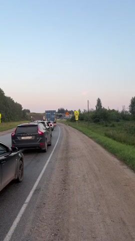 A traffic jam is visible on a rural road. Several cars are lined up on the right side of the road, facing away from the camera. A large blue truck is at the front of the line. In the background, there is construction work being done on what appears to be a bridge or overpass. There are also several large blue tanks or pipes visible near the construction site. Traffic signs indicating road work and a railway crossing are posted along the side of the road. The sky is clear and a soft pinkish hue suggests it is either sunrise or sunset. The road is paved on the left and dirt on the right. Trees and vegetation line the sides of the road.