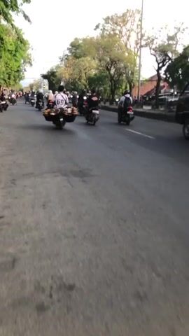 The media file depicts a busy street scene in Ngaban, East Java, with multiple motorcycles and scooters navigating the road. The subjects are primarily people riding motorcycles, many of whom are carrying goods. One prominent rider in the foreground wears a white shirt with a graphic on the back, and their motorcycle is laden with a large, multi-tiered rack of items, possibly food or merchandise. Other riders are also seen with various cargo.

The scene suggests an active commercial or logistical environment. The setting is an asphalt road with multiple lanes, bordered by trees and buildings on either side. The presence of overhead utility wires and streetlights indicates an urban or semi-urban area. The time of day appears to be late afternoon or early evening, as the sunlight is angled and casts shadows. The sky is partly cloudy, suggesting fair weather.

There is some visible text on the back of the white shirt worn by the prominent rider, appearing to be the number "64" in a stylized font. The overall mood is one of bustling activity and daily life. The location cues like the style of the trees and architecture are consistent with Southeast Asian settings, and specifically East Java as indicated. The overall impression is a vibrant street carrying out its daily functions.