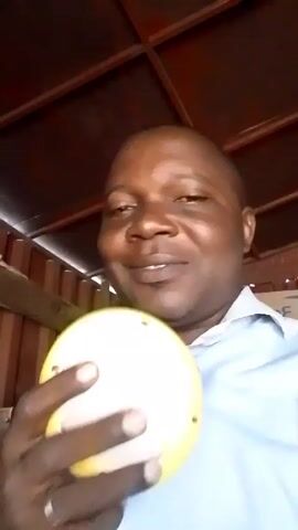 A man is smiling at the camera while holding a small, yellow-and-black object in his left hand. He is wearing a light blue shirt and a silver ring on his ring finger. The background features a ceiling with a dark red or brown panelled design. A wooden structure is visible behind him to the left. The man is close to the camera, focusing the shot on his face and the object in his hand.