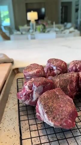 The video shows a close-up shot of raw, seasoned beef steaks arranged on a wire rack, which sits on a metal baking sheet. The steaks are thick-cut and appear to be seasoned with salt and pepper. The background is blurred, revealing a bright, modern kitchen or dining area with a large marble countertop. Further back, a dining table is set with plates and candles, and a television screen is visible. To the right of the beef, another metal baking sheet holds several raw chicken breasts, also seasoned. The camera pans slightly to the right, revealing more of the kitchen environment. The scene appears to be a preparation for a meal.