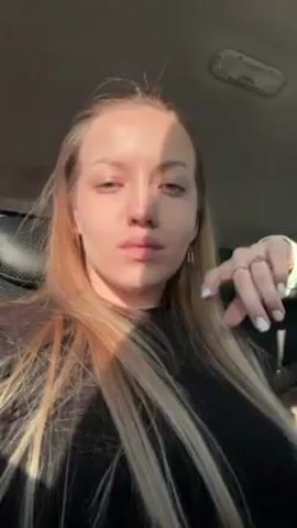 A young woman with long blonde hair is sitting in the backseat of a car. She is wearing a black top and several rings. The sun is shining through the car window onto her face. The car appears to be in motion. The interior of the car includes leather seats and a window.