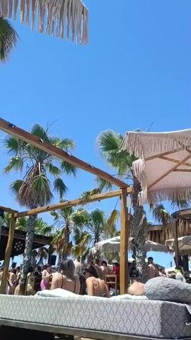 The video shows an outdoor scene on a sunny day, likely a beach or poolside bar. Several palm trees are visible, and there are wooden structures with thatched coverings. A crowd of people are gathered, some standing and talking, others seated. Some individuals appear to be dancing. The camera is positioned on a wooden platform, with a large, patterned cushion in the foreground. The sky is clear and blue.