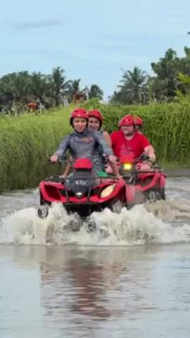 The video shows two red all-terrain vehicles (ATVs) driving through water. The first ATV has two people on it, a young male driving and a young female in the back. Both wear red helmets. The driver smiles and looks directly at the camera. The ATV has the lettering "KO4" on its front. The second ATV has at least two people, an adult male wearing a red shirt and helmet who is driving, with at least one other person behind him, also wearing a red helmet. Tall grass and palm trees are visible in the background. The sky is overcast.