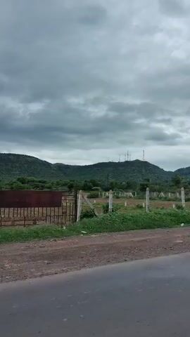 A cloudy sky looms over a rural landscape. In the foreground, a paved road runs horizontally across the bottom of the frame. Beyond the road is a dirt path, and further back, a fenced area with concrete posts and wire. Green vegetation and trees dot the landscape, leading up to rolling hills in the background. Several telecommunication towers are visible on the highest hill. On the right side of the frame, a small white building with "LADIES" painted on it stands next to a red-roofed structure. A man in a plaid shirt and dark pants stands near the white building, holding a pink object, possibly a ball. The scene appears to be a roadside rest area or an undeveloped plot of land.