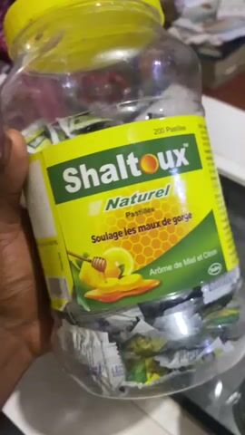 A person's hand, with dark skin, holds a large, clear plastic jar filled with individually wrapped cough drops. The jar has a bright yellow lid. The label on the jar is predominantly yellow and green, featuring the brand name "Shaltoux" in large, bold, white letters with a red outline. Below this, in smaller white text, it says "Naturel." Further down, the label displays "Pastilles" and "Soulage les maux de gorge," indicating that the product is for sore throats. An illustration of honey dripping from a honey dipper onto a slice of lemon and an orange slice is visible. The text "Arôme de Miel et Citron" (Honey and Lemon Flavor) is also present.

The label also indicates "200 Pastilles." The cough drops inside the jar are wrapped in silver and green foil, with some showing glimpses of yellow and blue. The background is blurred but suggests an indoor setting, possibly a home or shop, with stacks of paper or books visible behind the jar. The lighting is neutral, not suggesting a specific time of day. The overall impression is of a product being presented or examined. The location context provided is Jere South, Nigeria.
