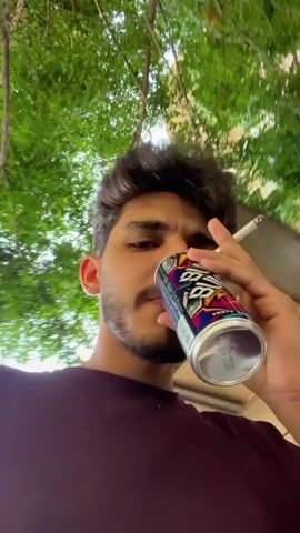 The video shows a young man holding a colorful can and a cigarette. He’s wearing a maroon shirt and standing outdoors under a tree with buildings visible in the background. He looks at the camera and seems to be filming himself. Another person wearing glasses and a black shirt walks by in the background, talking on a phone. The primary focus is on the man holding the can and cigarette.