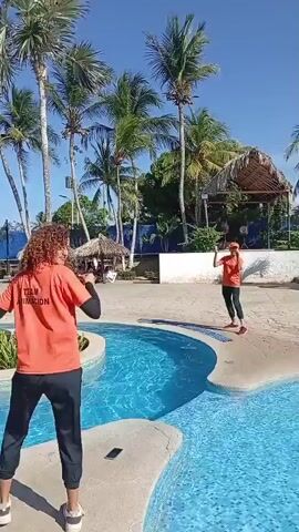 The video shows a sunny day at a resort with a large, winding pool surrounded by palm trees, thatched-roof structures, and greenery. Two people wearing orange "Team Animacion" shirts and dark pants dance to music near the pool, apparently leading an activity.  Other individuals are visible around the pool: some walking, some in the water, and others relaxing on the pool's edge. The pool has small, circular islands with plants in the middle, and there is an inflatable bouncy structure in the background. The overall atmosphere is relaxed and cheerful.