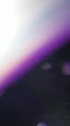 A DJ in a nightclub is the main subject of this video. The DJ, a young man with dark hair, is wearing a white jacket open in the front, revealing his bare chest and a necklace with a cross. He's using professional DJ equipment, including a mixer and turntables, which are visible in the foreground. The lighting in the club is dark with purple hues, creating a vibrant and energetic atmosphere. Several other people are around the DJ, some blurred and out of focus, suggesting they are dancing or moving around the club. Some of them are smiling and looking at the DJ. The background includes a mirrored wall, reflecting the lights and activity of the club. The DJ puts on headphones and interacts with the mixing equipment.