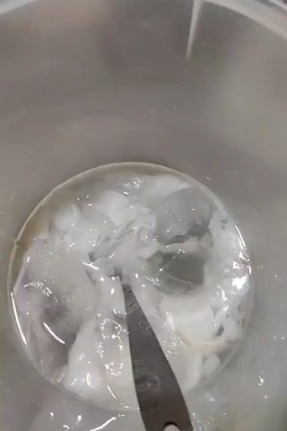 A close-up video shows a clear liquid with white, swirling substances inside a clear bucket. The contents are stirred with a flat metal spatula. The spatula disappears from view and the scene cuts to a light pink bucket containing a mixture of whitish liquid with what appears to be solids. A whisk is used to mix the substance. The scene cuts back to the clear bucket with the white swirling substance and a flat metal spatula mixing it.