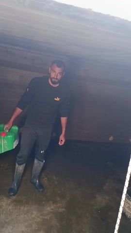 A man stands in a dimly lit, cavernous space with a low concrete ceiling. He is holding a green container with a red handle in his left hand. The floor is wet and appears to be made of concrete, with varying shades of brown and dark gray, suggesting possible staining or dampness. The man is wearing black rubber boots, black athletic pants, and a black long-sleeved Puma sweatshirt. He has dark hair and a beard, and he is looking directly at the camera with a serious expression. Part of a white net or mesh structure is visible in the lower right corner of the frame.