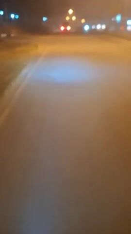 The video is shot from the perspective of someone riding a motorcycle at night on a paved road. The headlights of the motorcycle illuminate the road ahead, creating a bright white-blue glow that fades into the darkness. In the distance, the red taillights of a vehicle are visible, suggesting traffic ahead. Streetlights cast an orange hue on the surroundings, and there are blurry shapes that could be trees or buildings along the side of the road. The overall impression is one of movement through a dimly lit, possibly rural or suburban, environment at night.