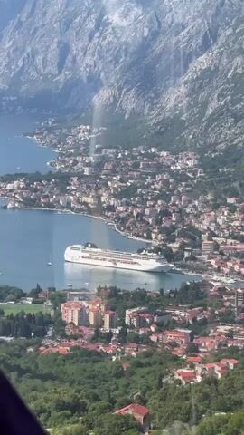 The video presents an aerial view of a bay city with a large cruise ship docked in the harbor. The city is nestled along the coastline at the base of a large mountain range. The urban area consists of a dense arrangement of buildings, many with red-tiled roofs, interspersed with patches of greenery. In the foreground, a hillside covered with trees provides an elevated perspective of the city below. The large cruise ship, predominantly white with a dark blue hull, is a prominent feature in the bay. The sky is mostly clear, allowing for sharp detailing in the cityscape and mountainous backdrop.