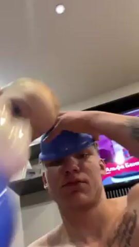 A shirtless man with a blue cap and tattoos takes a selfie video with a blue plastic device with a white hippopotamus figure attached to the top. The device is humming. The man appears to be inside a house. There is a television visible in the background.